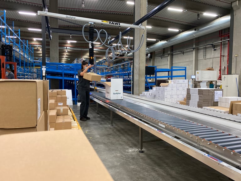 Employee at Qualiphar using a TAWI vacuum lifter to move heavy boxes into packaging boxes on a conveyor