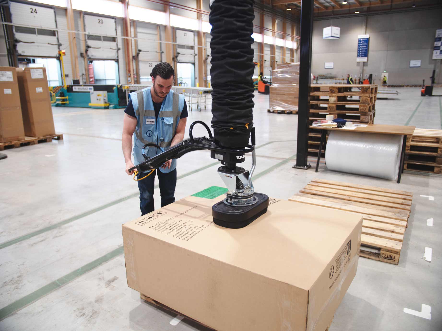 Logistics warehouse worker at Kuehne Nagel France using TAWI vacuum lifter to palletise boxes