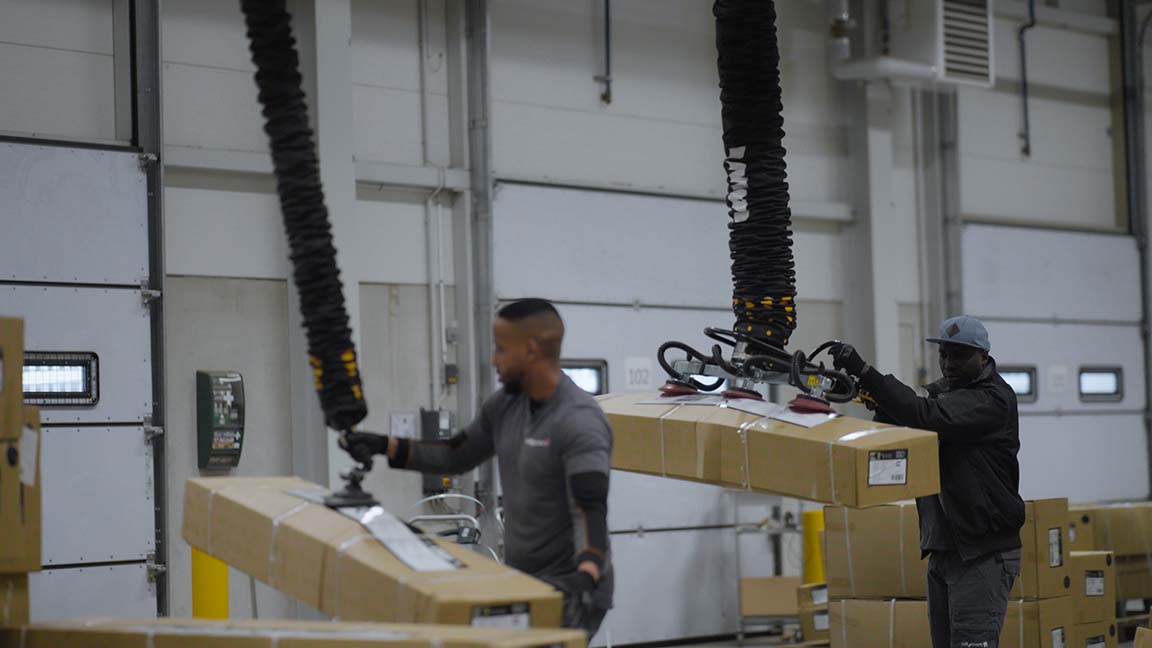 Two men lifting bulky packages with handheld vacuum lifters at Jollyroom warehouse